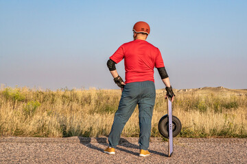 mature senior male with one-wheeled electric skateboard on a prairie road, late summer scenery with...