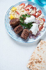 Grilled kofta kebabs with couscous, red tomatoes, white yogurt and flatbread, top view on a white stone background, vertical shot with space