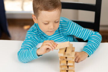 A cute seven-year-old child boy playing Jenga at home at a white wooden table against the background of a light window. Selective focus. Close-up. Portrait