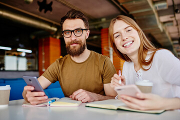 Portrait of cheerful male and female students smiling at camera during together learning in coffee shop, happy Caucasian bloggers posing during daytime meeting for discussing mobile phoning via app