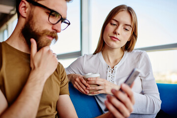 Millennial male blogger talking about idea for text publication with attractive female friend, Caucasian man and woman using mobile phone for browsing website connecting to 4g wireless internet