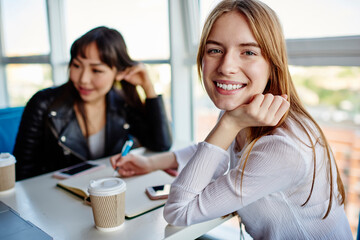 Portrait of cheerful Caucasian student smiling at camera while collaborate with colleague on informative notes from education textbook, happy diverse women preparing to university exams togetherness