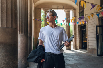 A young student walks in the courtyard of the university with a folder in his hands. Confident young man in glasses with a laptop in his hands.