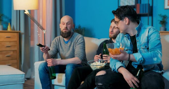 Three best friends spend evening together in front of TV screen drinking beer eating chips and popcorn bearded man switches channels with remote control looking for show interesting movie new series
