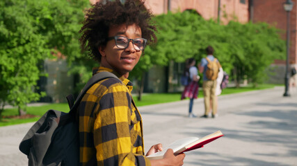 Afro-american male student reading book walking outdoors and looking back at camera