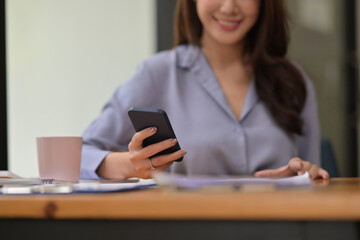 Cropped image of a young woman is using her smartphone while sitting at the wooden working desk.