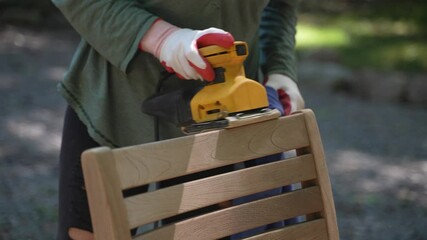 Closeup detail of woman holding electric sander sanding a teak chair outside with dust.