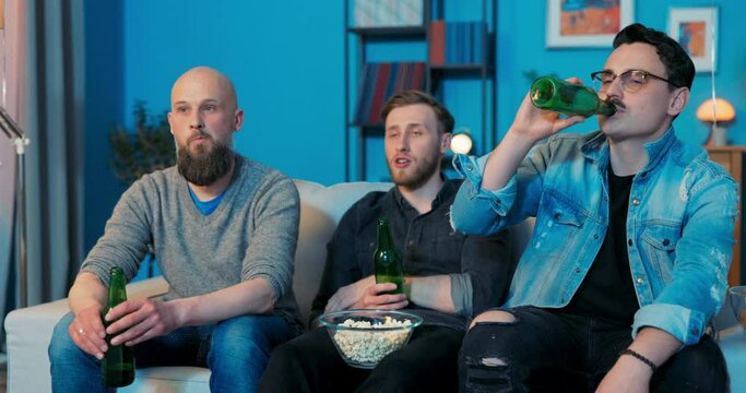 Three buddies are sitting on sofa in living room in evening watching their favorite TV series movie drinking beer, eating snacks from bowl, commenting together on action on TV, staring at screen