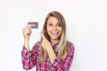 A young smiling blonde woman in a red plaid shirt holds a plastic credit card in her hand and points at it with her finger isolated on a white background. Shopping, payment for purchases, promotion