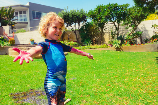 Blond boy in a summer costumes standing on green grass lawn with opened pink painted hands