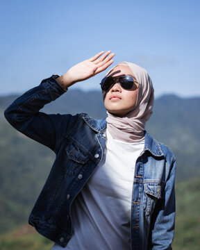 Hijabi woman in denim jacket wearing sunglasses blocking sunlight with her hand