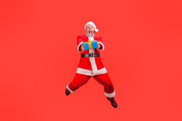 Positive elderly man with gray beard wearing santa claus costume jumping high with wrapped present box, giving gift for Christmas, looking at camera. Indoor studio shot isolated on red background.