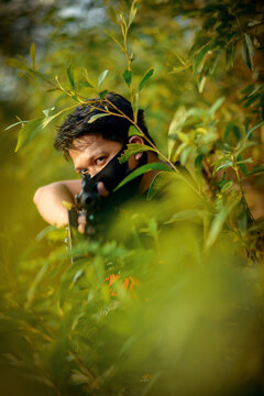 Man with facemask holding gun standing behind green tree