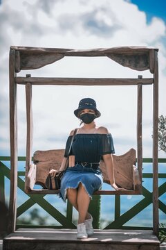 Woman in leather facemask and gray hat sitting on wooden swing outdoor