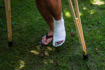 Young man in white plaster cast with broken leg and crutches walks on lawn. Broken leg. Close-up....