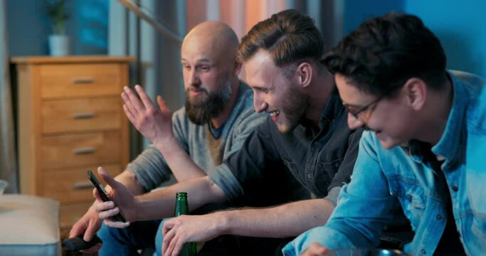 Three men are relaxing on the living room couch in the evening in front of the TV screen, sipping a beer, talking, the guy shows them on his phone a picture, photo, meme, laugh, joy, smile