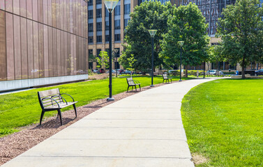 Scenery of bench placed along walkway in green public park, lush grass around outdoor garden for recreation and rest in sunny day. Spring and summer lifestyle with nature environment. Empty landscape.