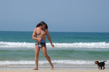 man gay walking in beach clothes, hat and sunglasses, on the sand with his little black dog, in the background the blue sea and its clear on a summer day
