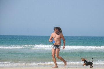 man gay walking in beach clothes, hat and sunglasses, on the sand with his little black dog, in the background the blue sea and its clear on a summer day
