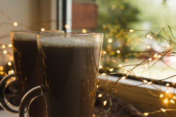 Close-up of couple cocoa drinks in glasses. Hot chocolate on sil. Led lights garland on background.