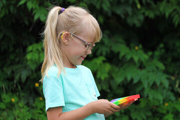 a blonde little girl playing with an anti-stress multi-colored pop toy on the street