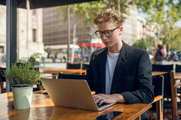A young businessman in a suit sits on the terrace of a cafe and works on a laptop. In a big city, a...