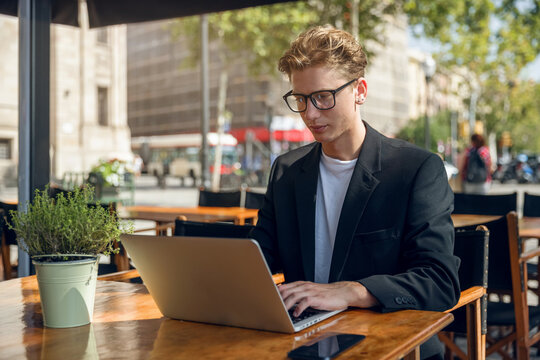 A young businessman in a suit sits on the terrace of a cafe and works on a laptop. In a big city, a business man sits at a table on the terrace and communicates via a laptop.