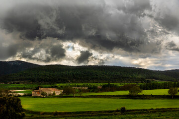 Obraz na płótnie Canvas Farm in a green meadow in a day with white and gray clouds and sun rays with mountains.