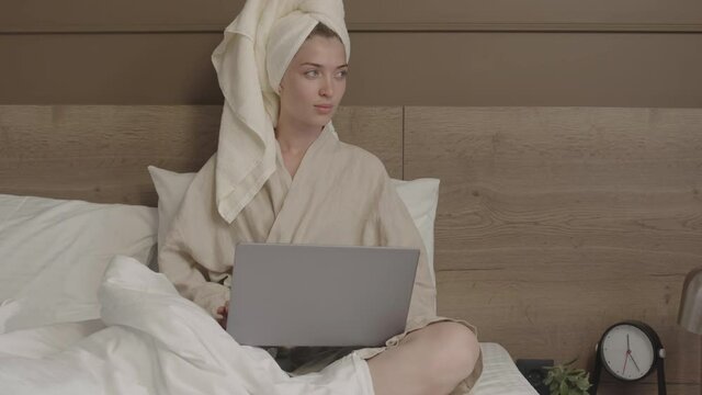 Medium shot of young Caucasian woman after bath with towel on her head working on laptop computer sitting in bed