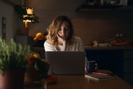 Bored woman browsing laptop in evening in kitchen