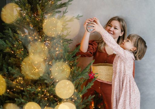 Cheerful girl with mother hanging bauble on green tree