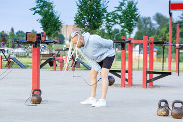 A man with an interesting braid hairstyle in a blue hoodie works out early in the morning on the street and makes swings with a kettlebell.