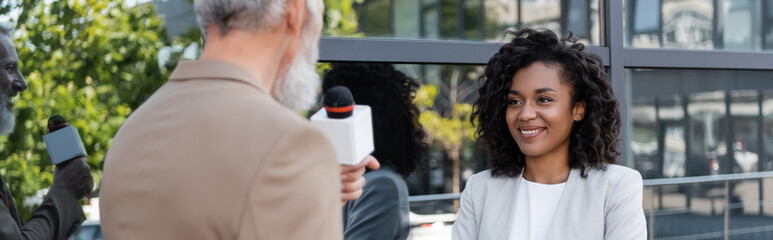 blurred and bearded reporter with microphone talking near smiling african american businesswoman,...