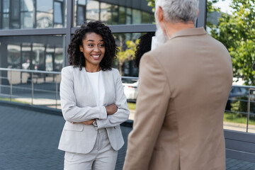 smiling african american businesswoman standing with crossed arms near blurred reporter