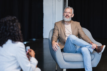 Fototapeta premium blurred african american journalist talking with confident businessman sitting in armchair during interview
