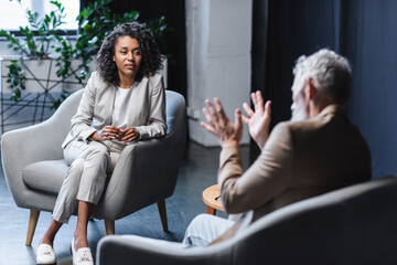 curly african american journalist looking at blurred businessman sitting in armchair and gesturing...