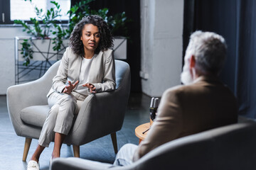 curly african american journalist gesturing while talking with blurred businessman sitting in...