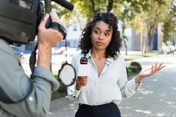 curly african american journalist with microphone pointing with hand while doing reportage near blurred cameraman - obrazy, fototapety, plakaty