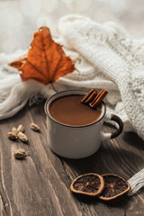 Mug of hot chocolate or cocoa with cinnamon sticks on a wooden table against a background of autumn leaves.Warm scarf and cozy autumn and winter concept. 