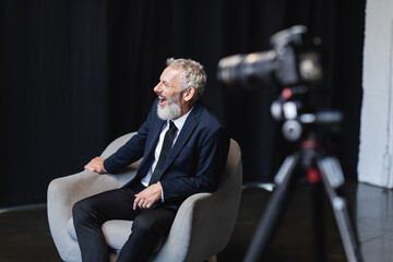 cheerful businessman in suit laughing while sitting in armchair during interview near blurred...