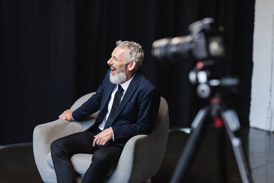cheerful businessman in suit laughing while sitting in armchair during interview near blurred digital camera on tripod