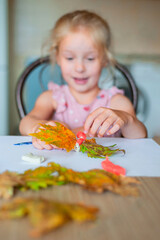 Little smiling girl looking at her autumn craft A little girl made a figurine of plasticine and autumn leaves on the table at her home. Homemade autumn creativity