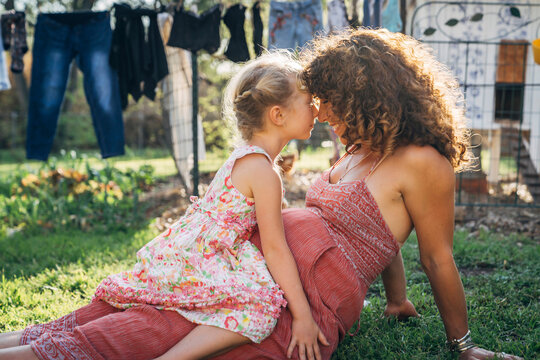 Girl sitting on pregnant mom's lap outside on lawn touching noses 