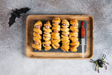 Halloween mummy hot dogs. Sausages with eyes wrapped in dough in wooden tray on concrete background. Crazy funny Halloween food. Top view, flat lay.
