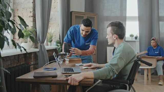 Medium slowmo shot of member of volunteer team helping young man with disability in wheelchair to set up wi-fi router in his apartment