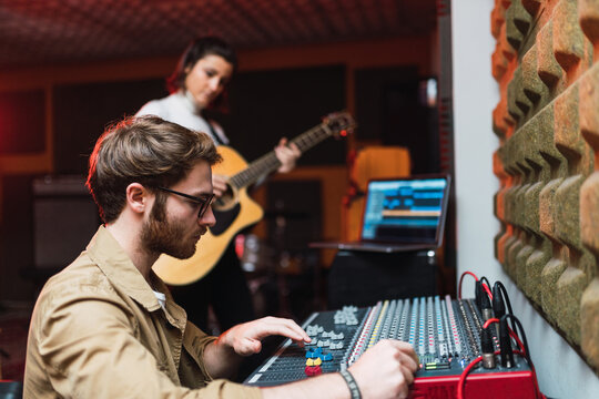 Woman playing on guitar near man using equalizer in studio
