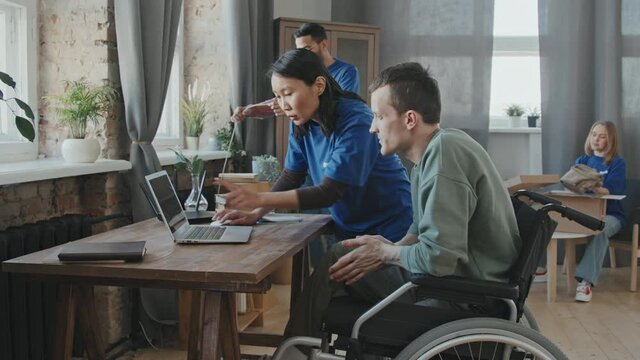 Tracking slowmo shot of multi-ethnic team of volunteers explaining young man with disability in wheelchair how to use internet on his computer while setting up wi-fi router in his apartment