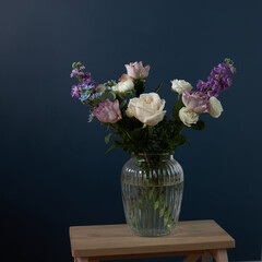 Bouquet of hackelia velutina, purple and white roses, small tea roses, matthiola incana and blue iris in glass vase . Dark blue wall