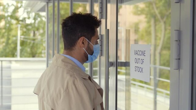 Young Man In Mask Reading Vaccine Out Of Stock Sign On Hospital Door