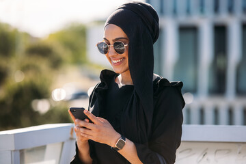 Happy Muslim woman in stylish outfit using cellphone